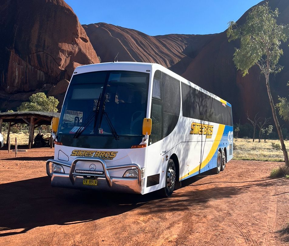 Coach parked in front of Uluru rock formation, white, blue, and yellow accents.