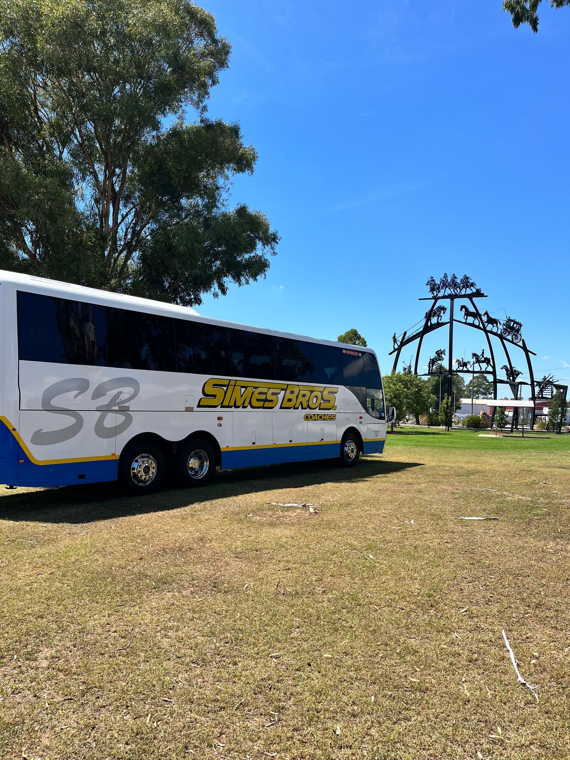 White and blue tour bus parked on grass, blue sky, tree, and metal sculpture in the background.