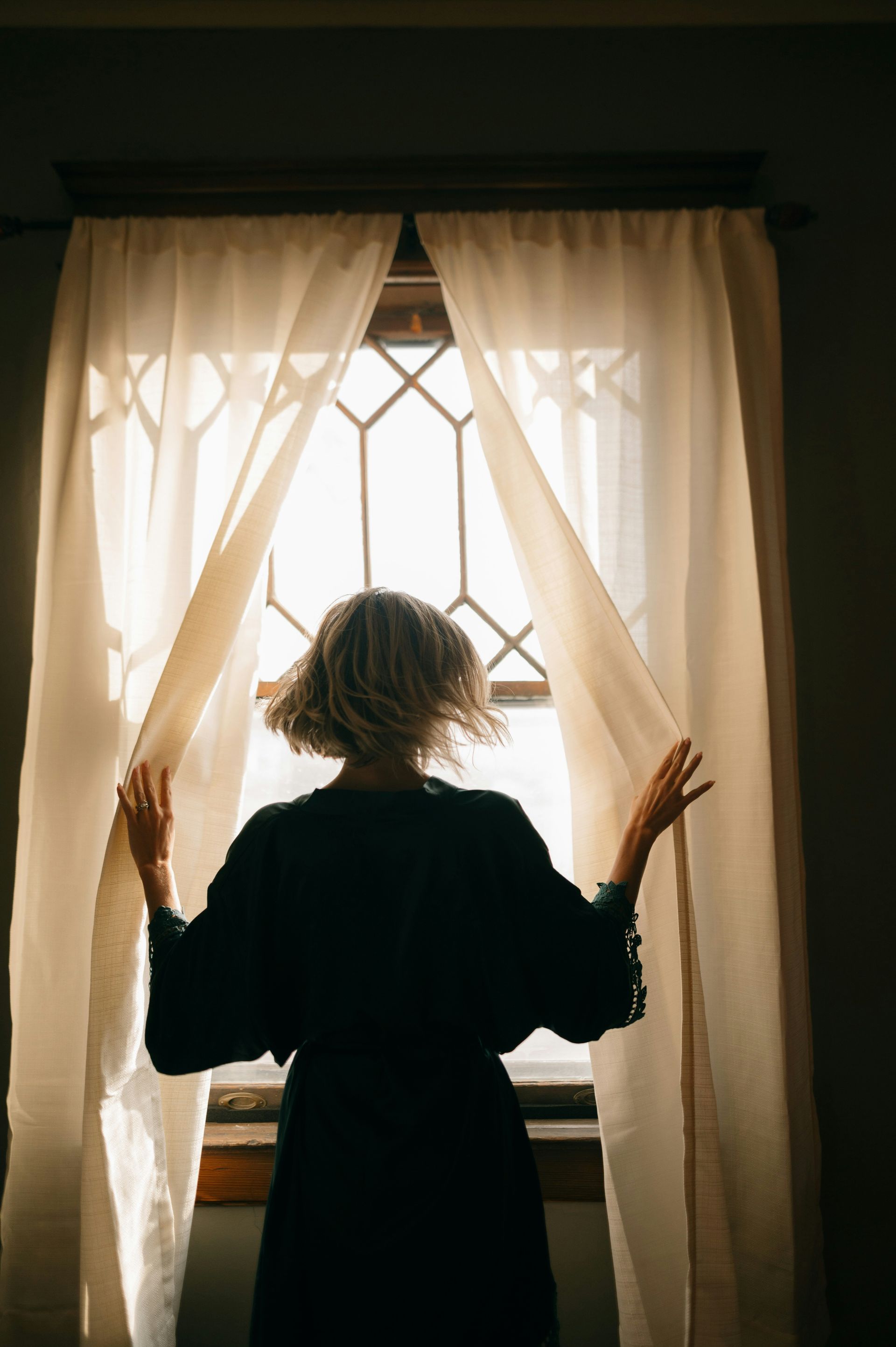 Woman looking out a window as light comes through the curtains