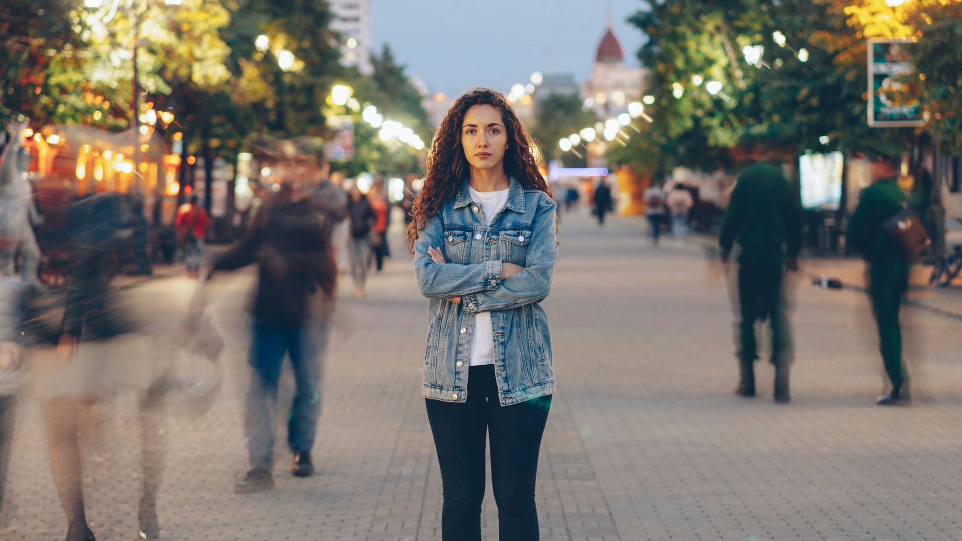 A woman standing with arms crossed on a crowded street, looking guarded while people move past her in a blur.