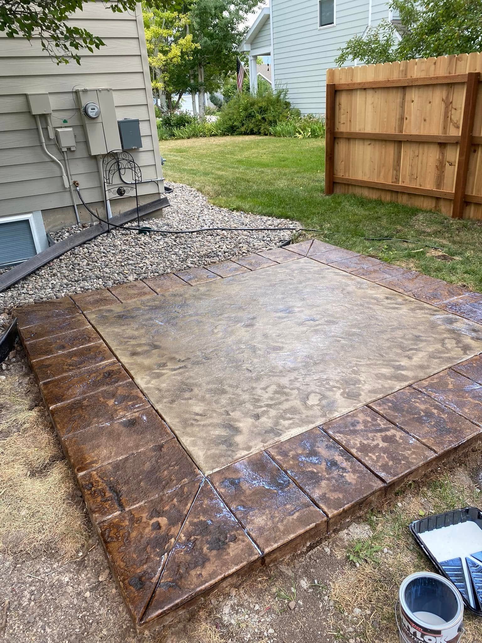 Concrete patio slab bordered by wet brown pavers beside a house and wooden fence in a backyard