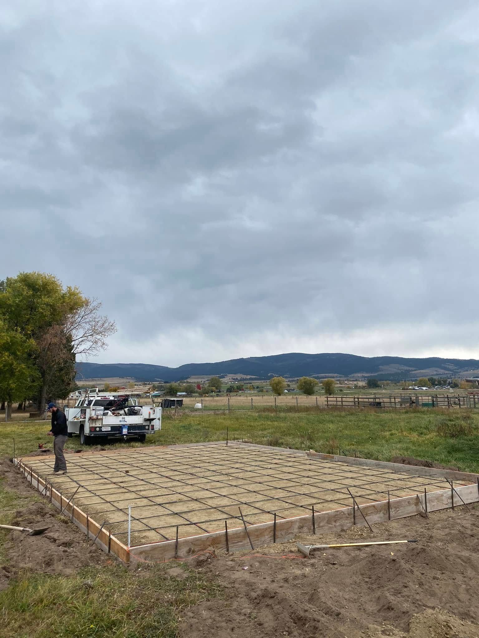 Construction foundation site with rebar grid, parked truck, and cloudy sky over open field