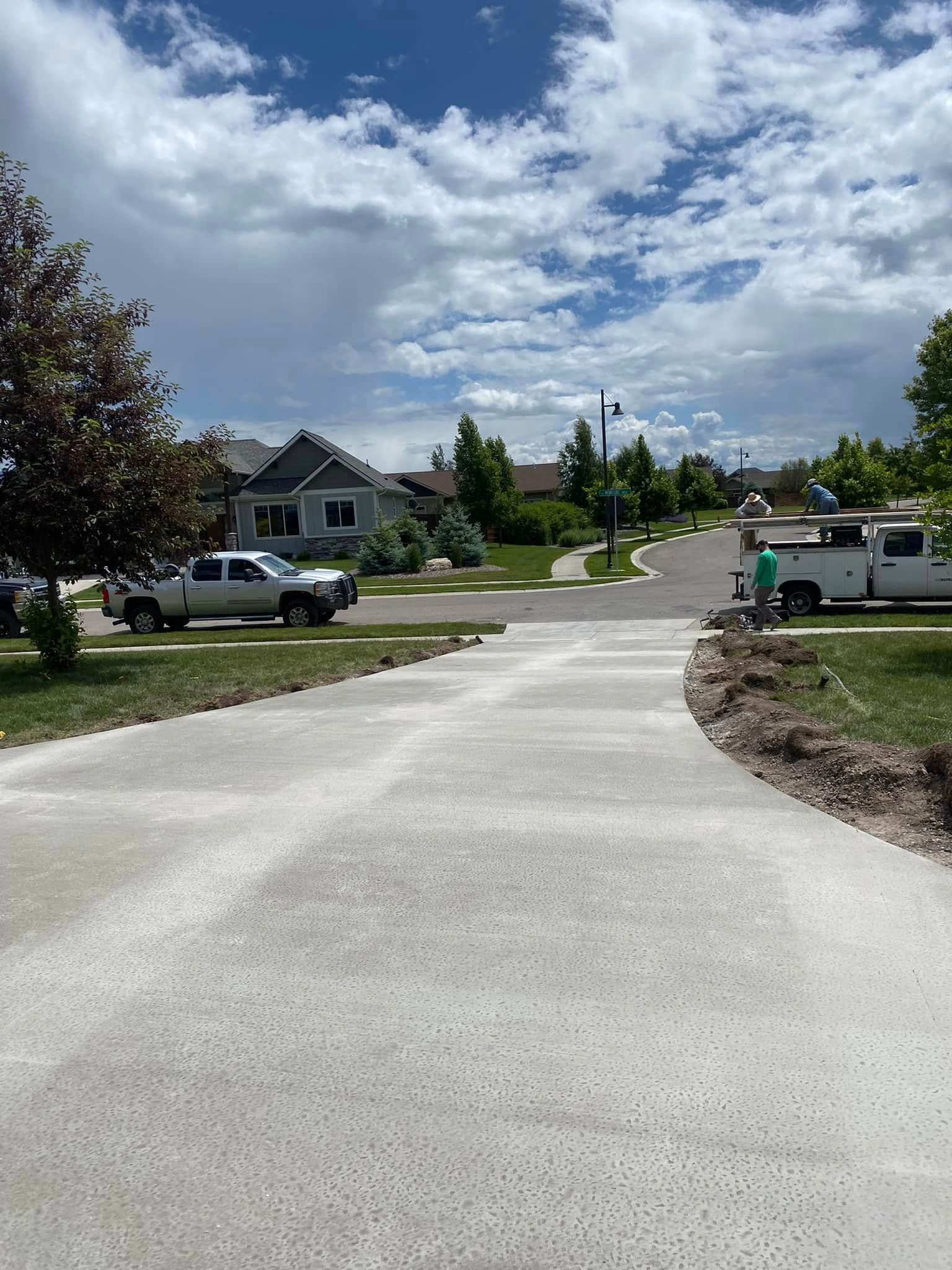 Curving paved driveway in a suburban neighborhood under a cloudy sky, with parked vehicles and houses nearby