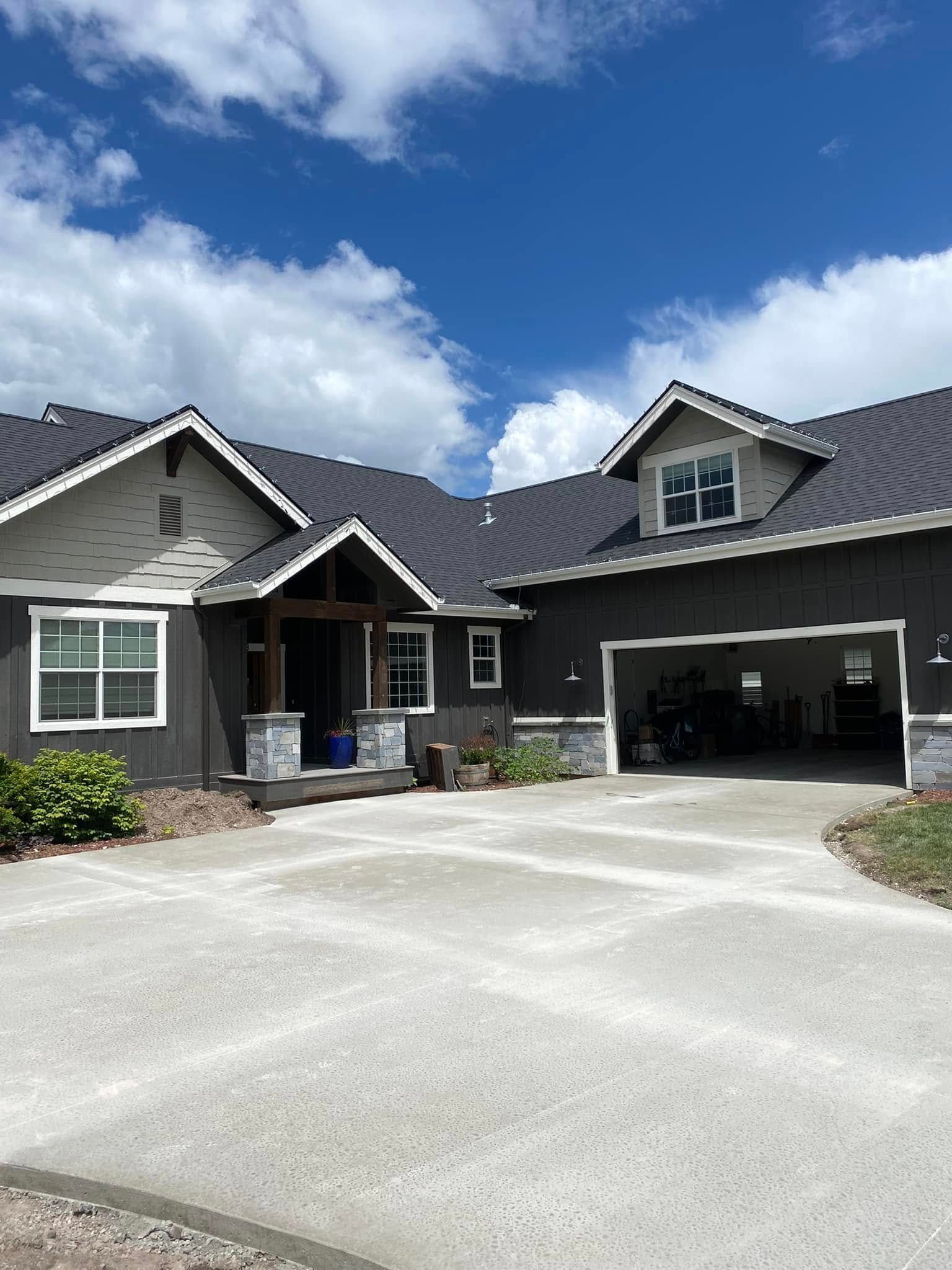Suburban house with dark roof, stone facade, and wide driveway under a partly cloudy sky