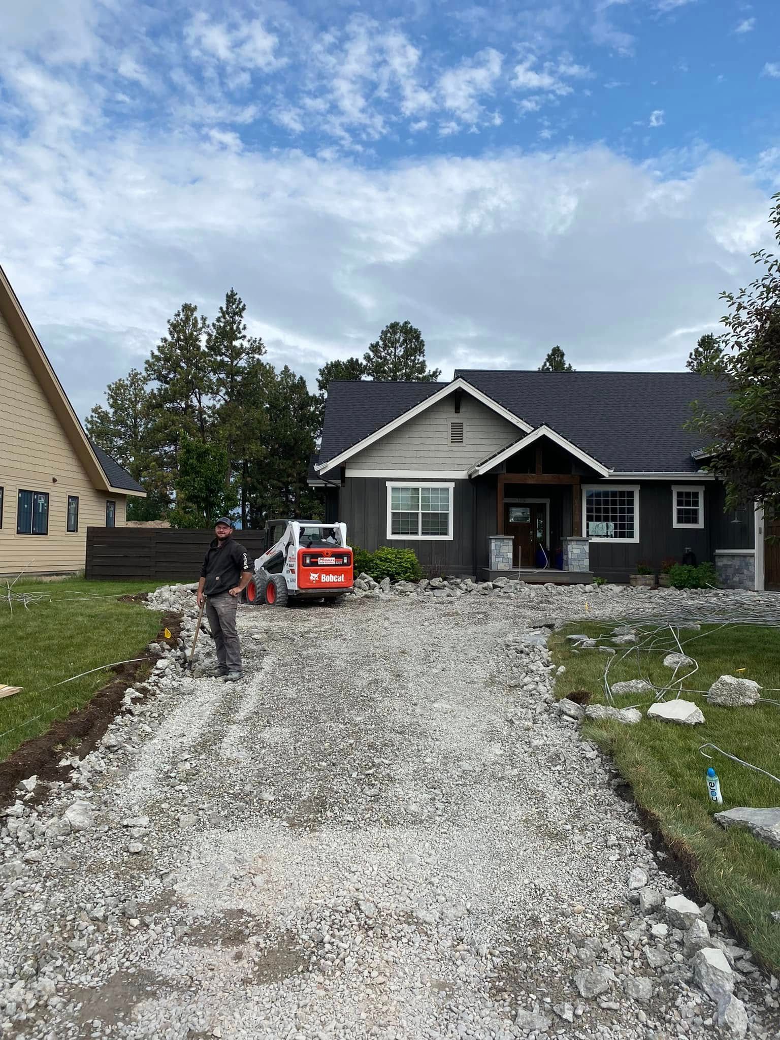 Gravel driveway under construction leading to a gray house, with a worker and orange equipment.