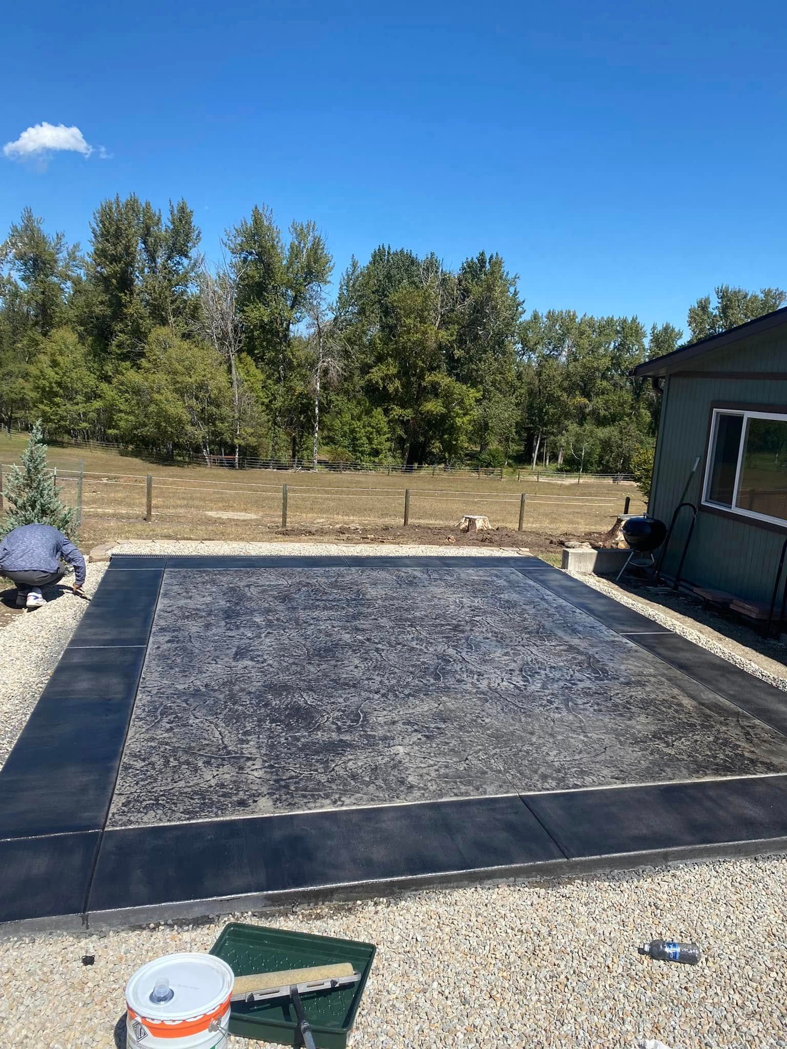 Backyard with a rectangular gravel and paver patio under a blue sky, bordered by trees and a house.
