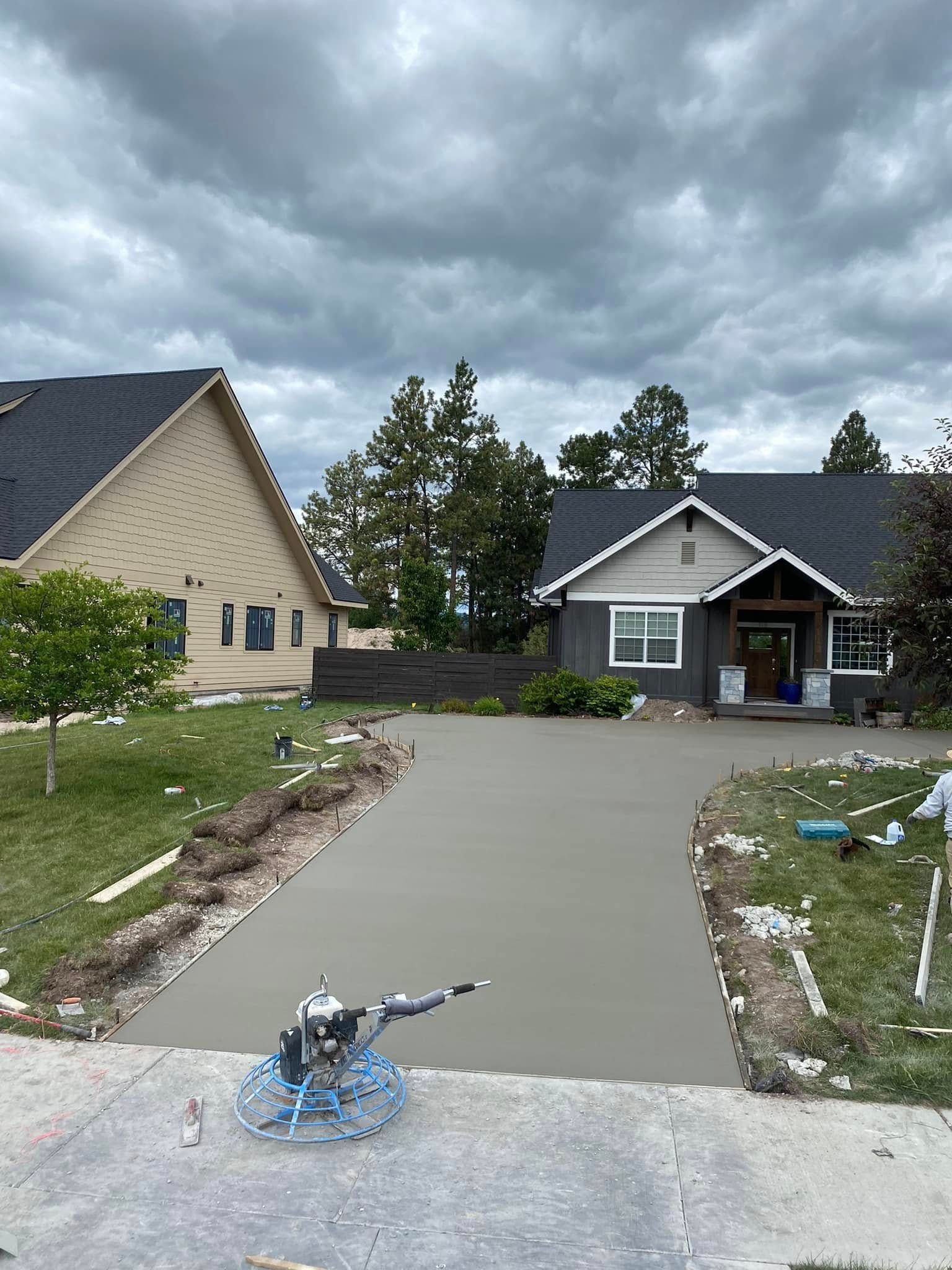 Fresh concrete driveway between two houses under a cloudy sky, with a hose lying on the wet slab.