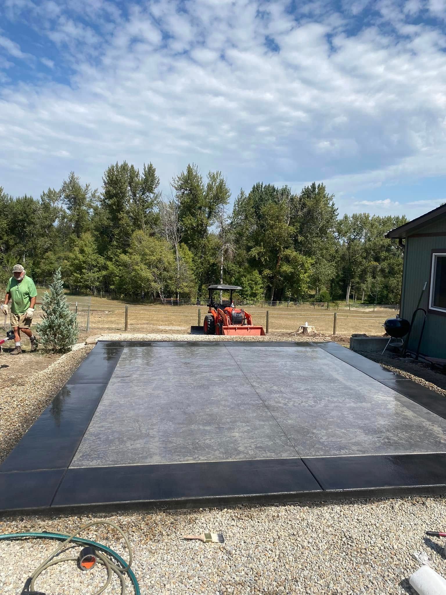 Outdoor patio under construction with gray paver base, black edging, gravel, trees, and a red compacting machine.