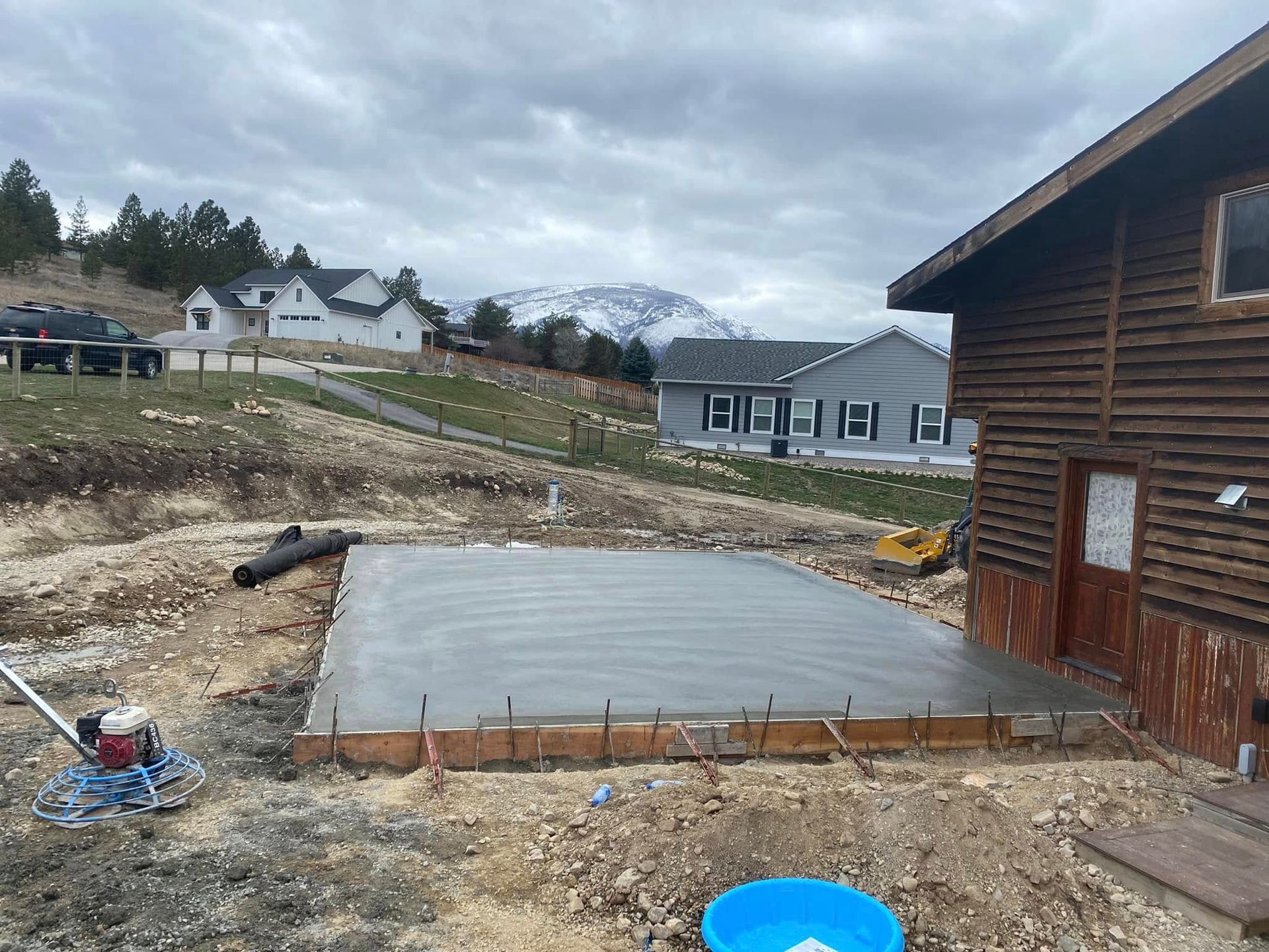 Concrete slab foundation beside a log cabin, with houses in the background under a cloudy sky.