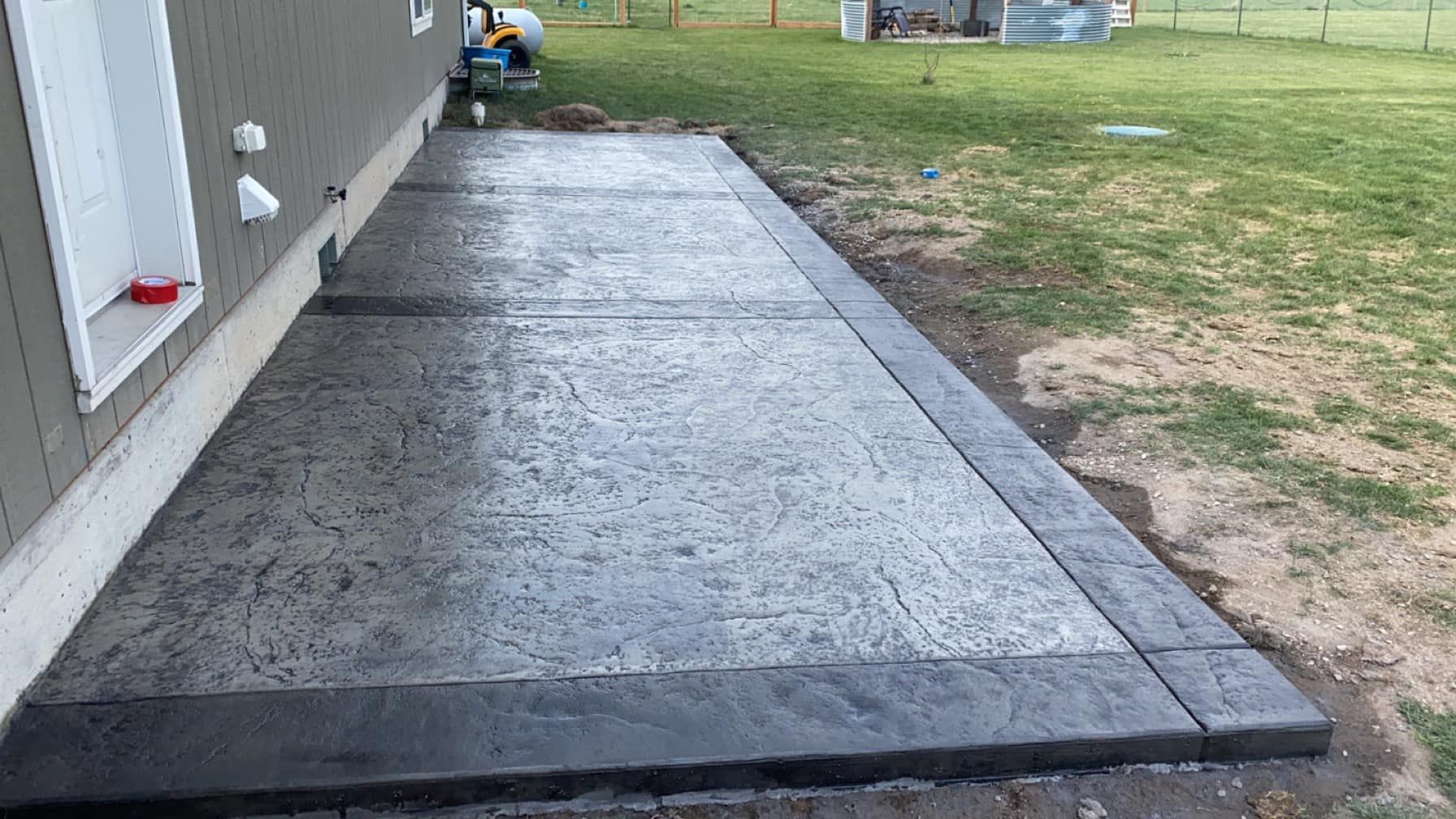 Concrete walkway beside a house, with a raised dark border and a grassy yard in the background.