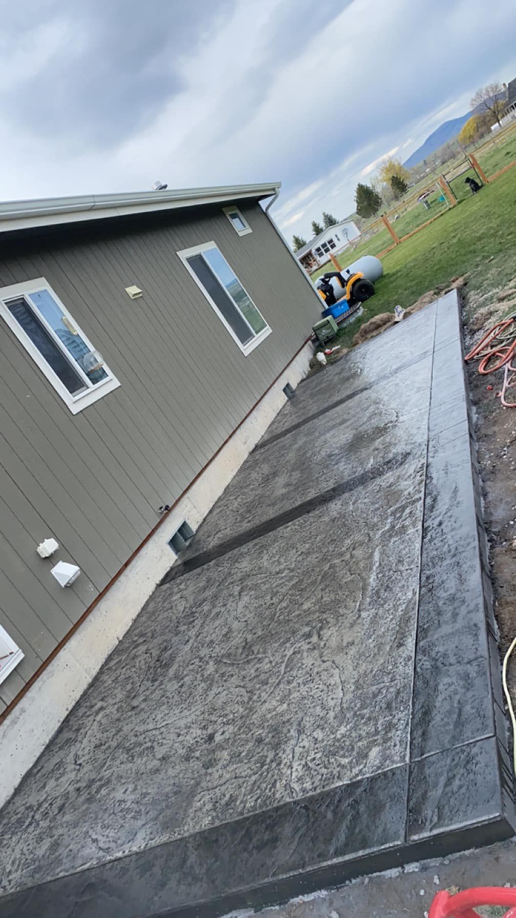 Tilted view of a gray house side yard with a concrete walkway and a worker in a green vest near the fence