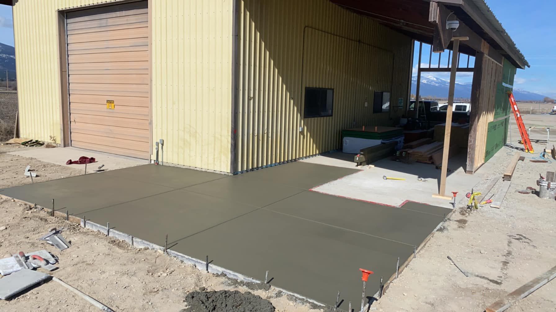 Fresh concrete slab and ramp beside a tan building under construction, with posts and tools nearby