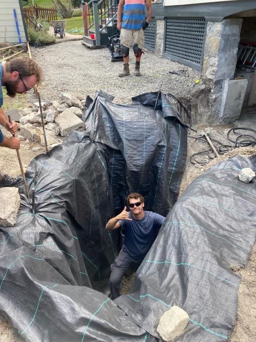 Man in sunglasses climbing out of a deep plastic-lined trench at a construction site, with workers nearby