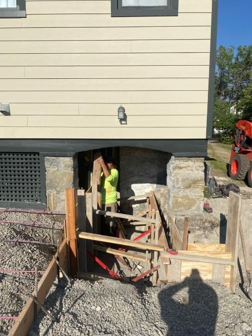 Worker in neon vest at a basement foundation opening under a house, with wooden supports and gravel.