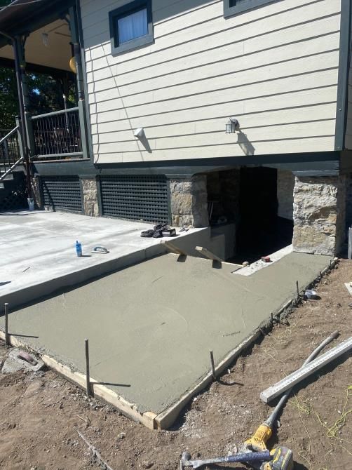 Concrete patio under construction beside house with basement doorway, wooden forms, and tools on dirt.
