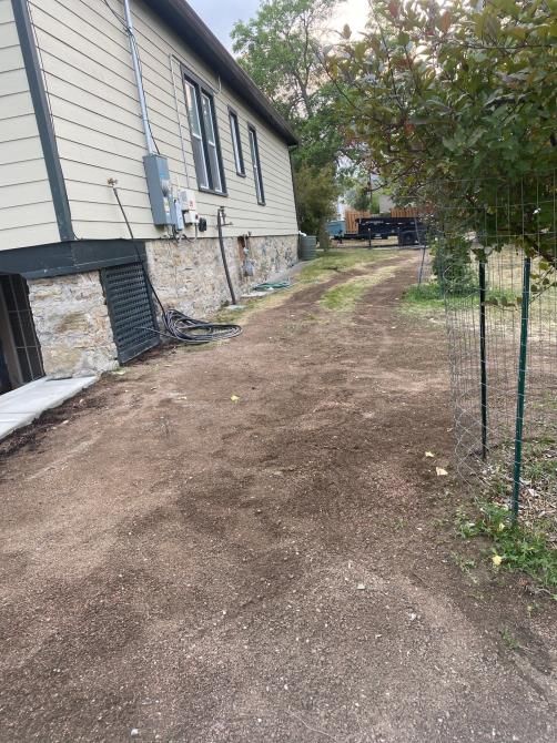 Side yard beside a house with a dirt path, stone foundation, and chain-link fence under trees