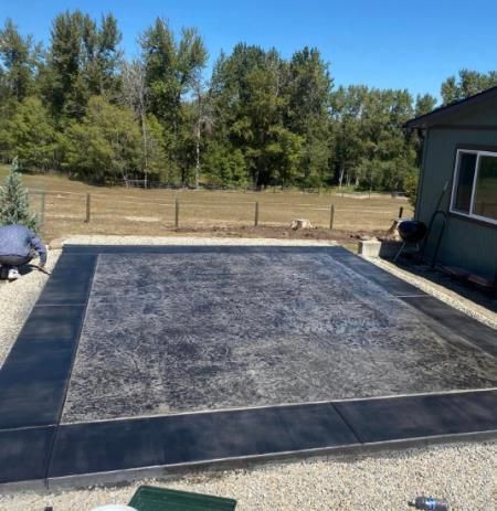 Backyard with a large rectangular black patio or deck under construction, bordered by gravel and trees.