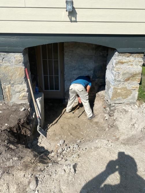 Worker shoveling dirt under a house porch opening beside a basement door and stone foundation.