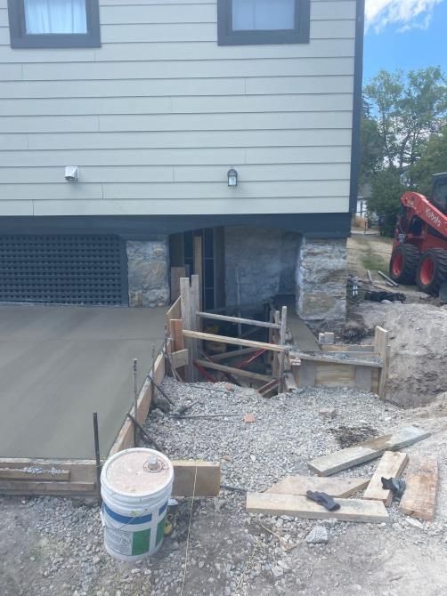 House foundation excavation with gravel, wooden supports, and a red machine beside the driveway
