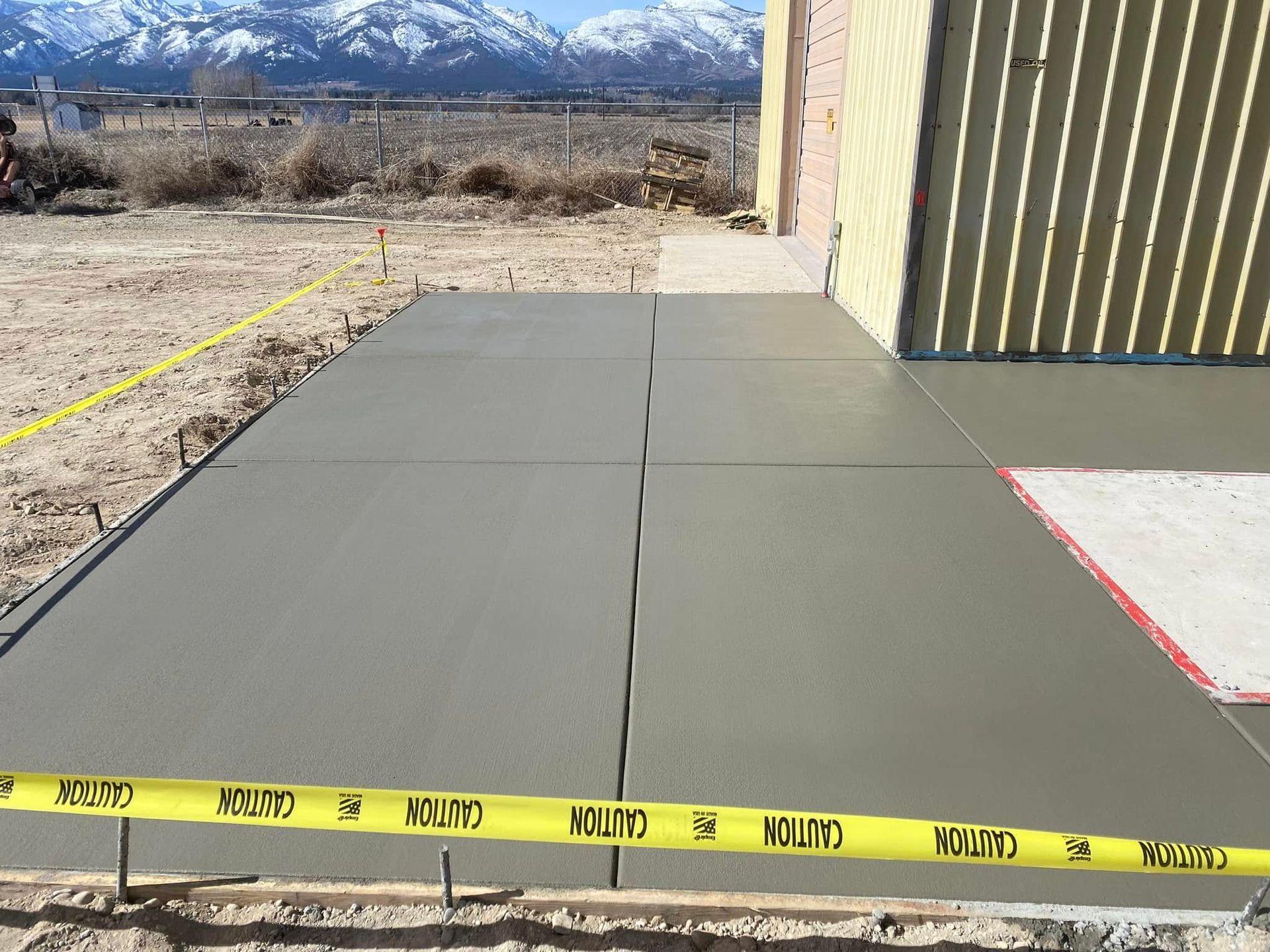 Fresh gray concrete walkway beside a tan building, marked by yellow caution tape, with mountains in the background