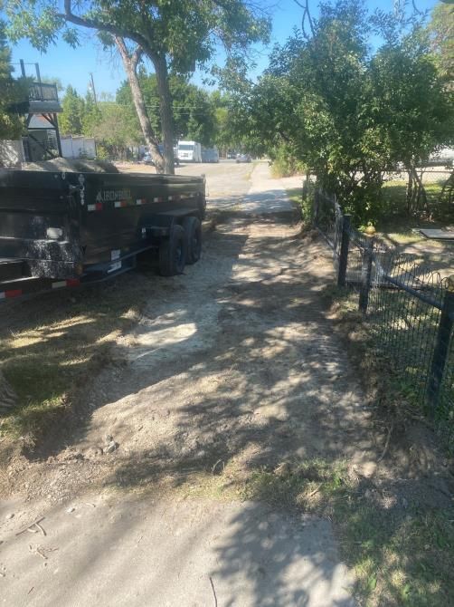 Dirt driveway beside a black pickup truck and chain-link fence under trees on a sunny day