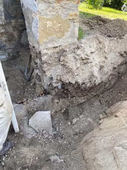 Excavation around a stone foundation corner with exposed soil and rubble beside a grassy yard