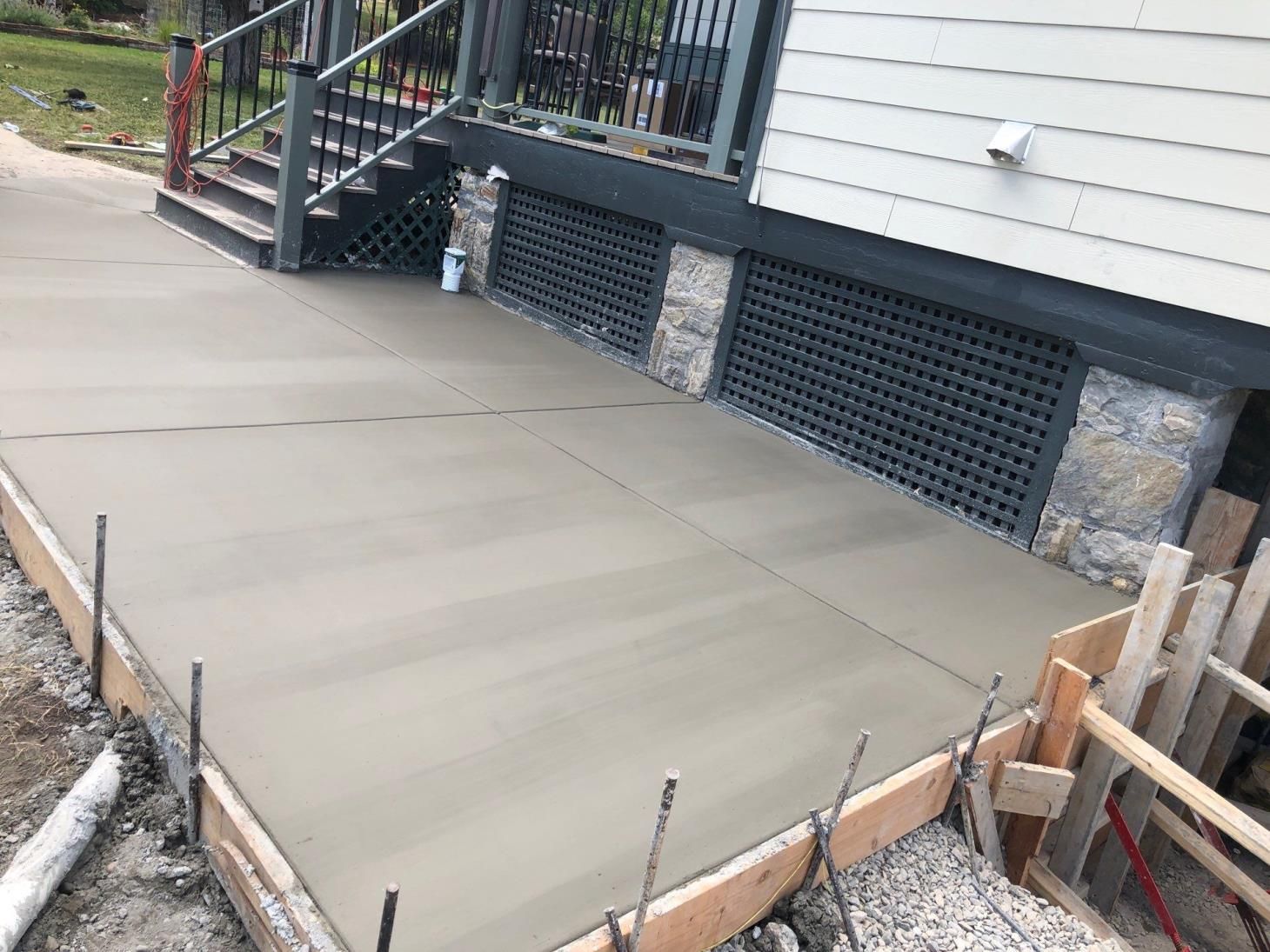 Freshly poured concrete patio beside a house, with steps, railing, and foundation vents.