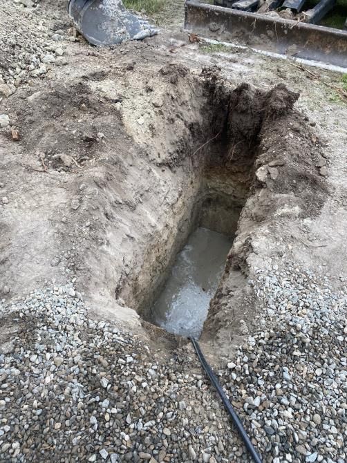 A deep trench dug in a gravel lot, exposing a narrow hole with muddy water at the bottom.