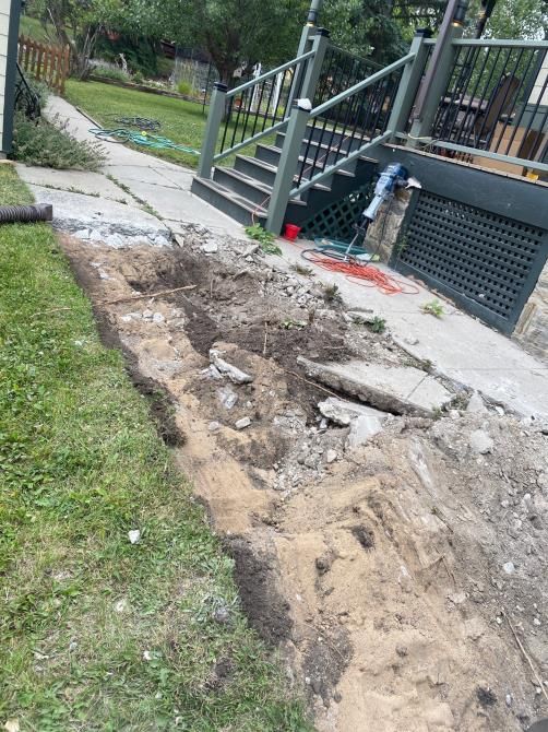 Damaged walkway with exposed dirt and broken concrete beside a porch and stairs