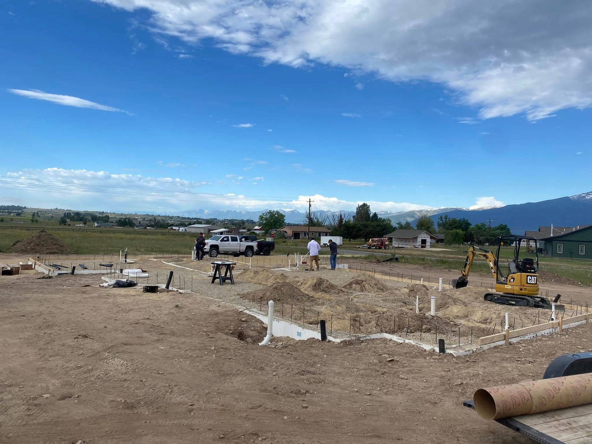 Construction site with foundation forms, workers, trucks, and a blue sky over open land
