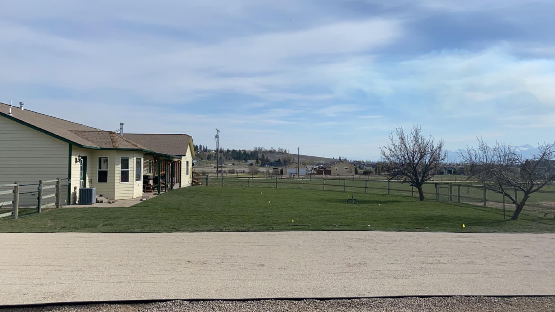 Wide open field beside a beige house and dirt road under a cloudy sky.
