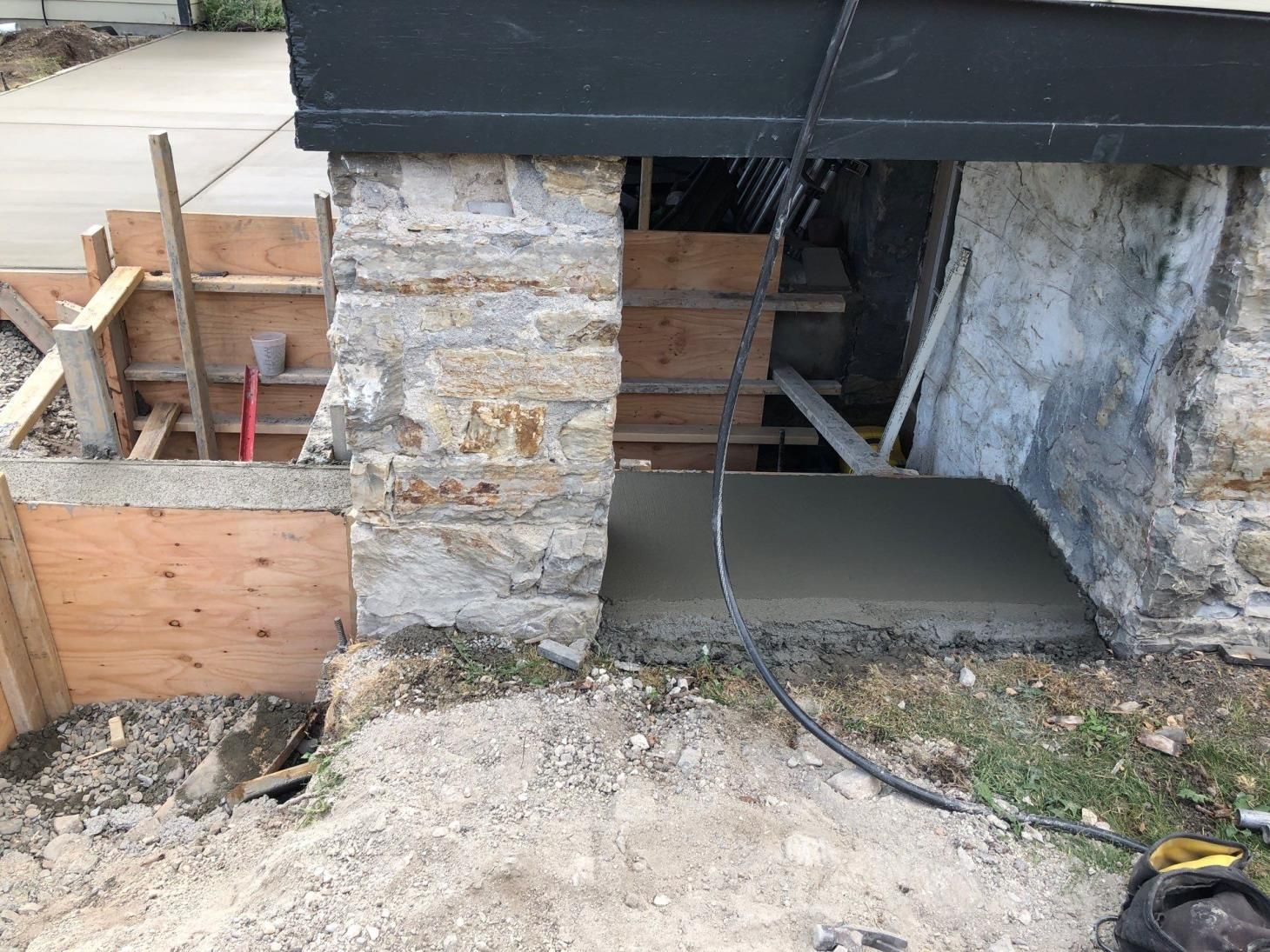 House foundation under construction with wooden forms, stone wall, and concrete slab poured around a support post
