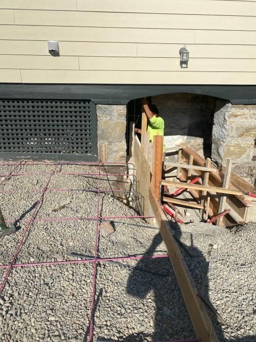 Worker in neon shirt in a basement excavation beside a house, with gravel, lumber, and support posts.