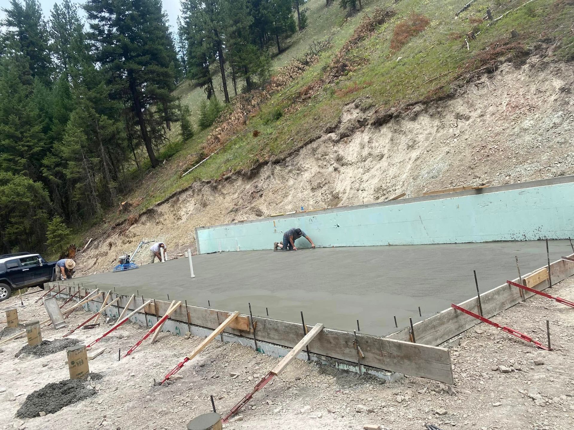 Fresh concrete foundation poured beside a hillside, with rebar, forms, and workers visible.