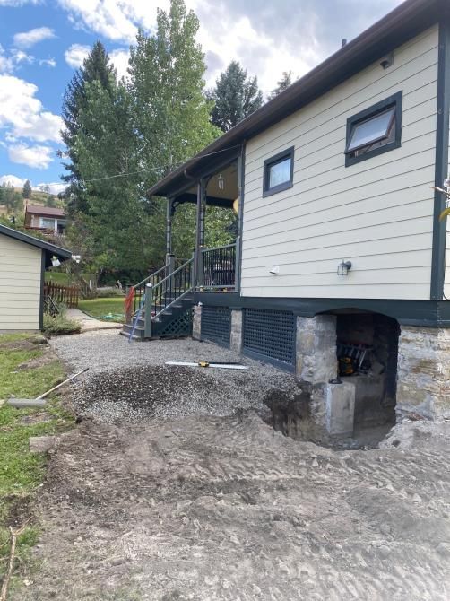 House raised on stilts with unfinished dirt slope and stairs beside a porch, under a partly cloudy sky