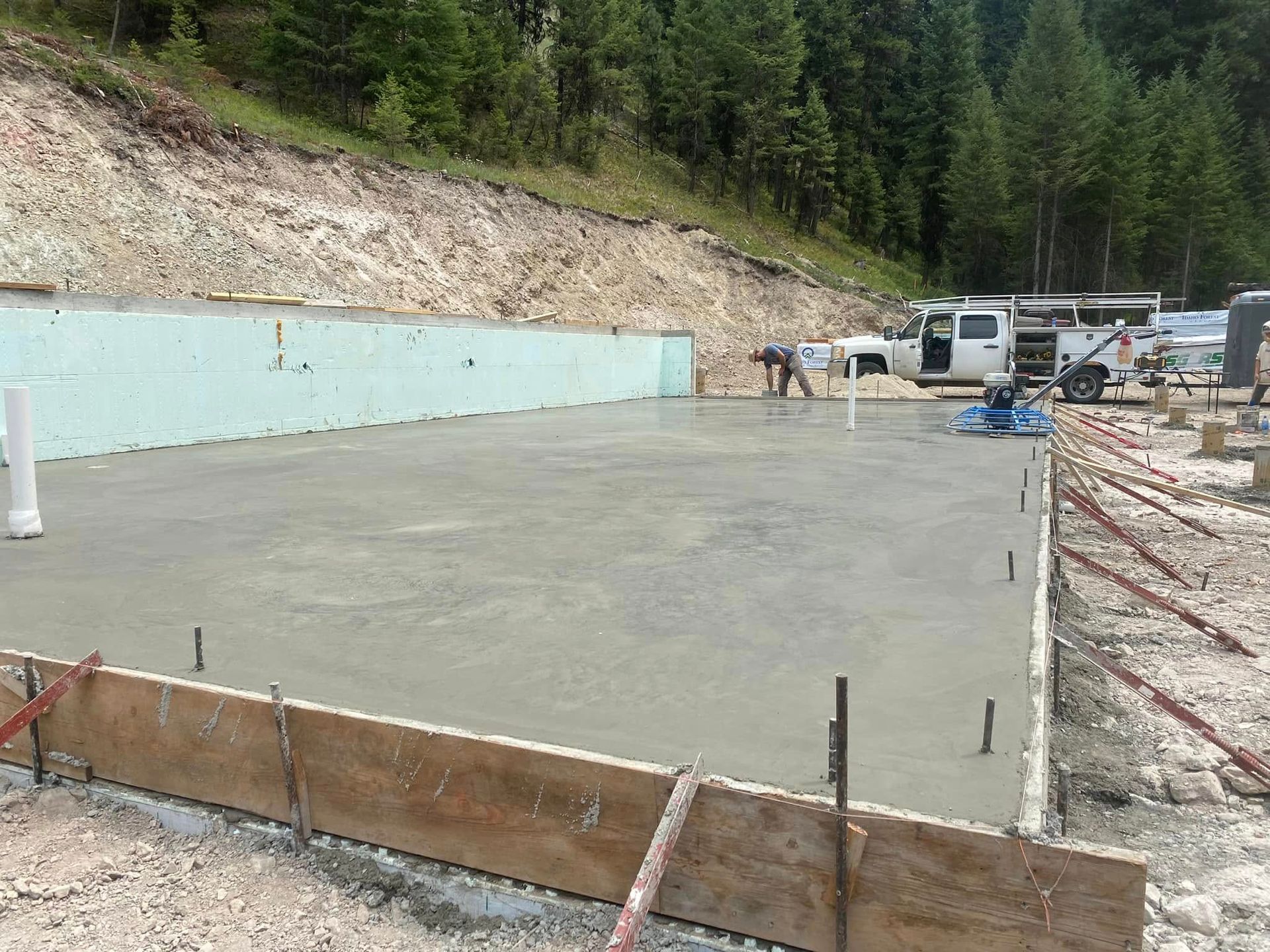 Fresh concrete slab at a construction site beside a forested hillside, with parked vehicles and forms around it