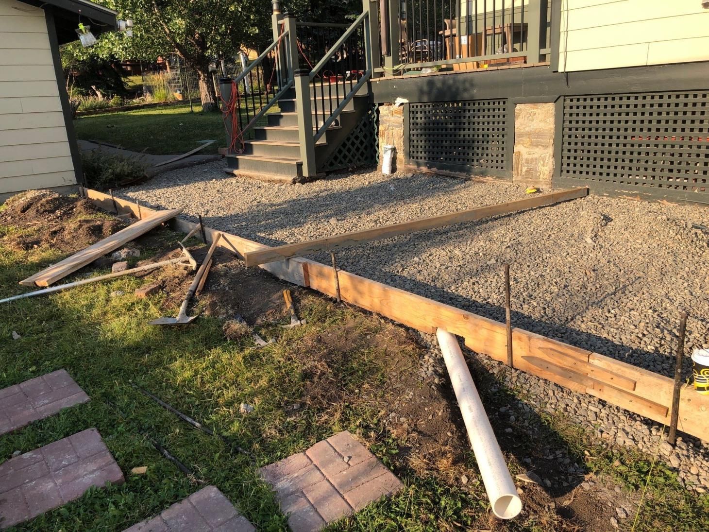 Front yard construction with gravel slope, wooden retaining edge, stairs, and a loose white pipe on the grass.