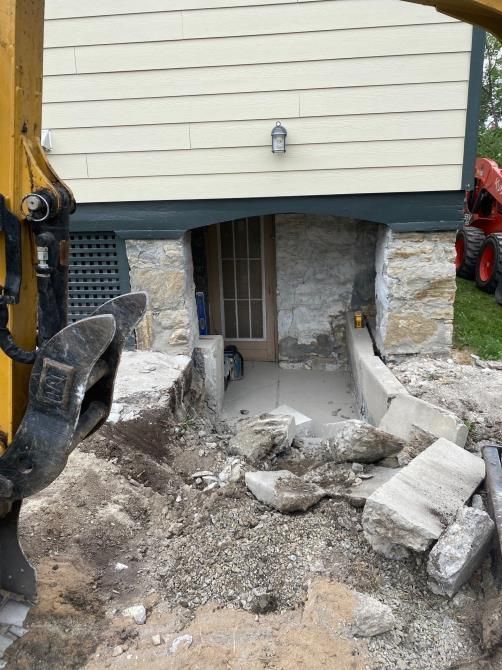 Excavator digging out a basement foundation beneath a house, with rubble and stone blocks around the opening