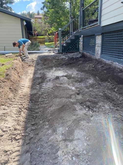 Worker leveling dirt beside a house and porch, with a narrow path area under construction.