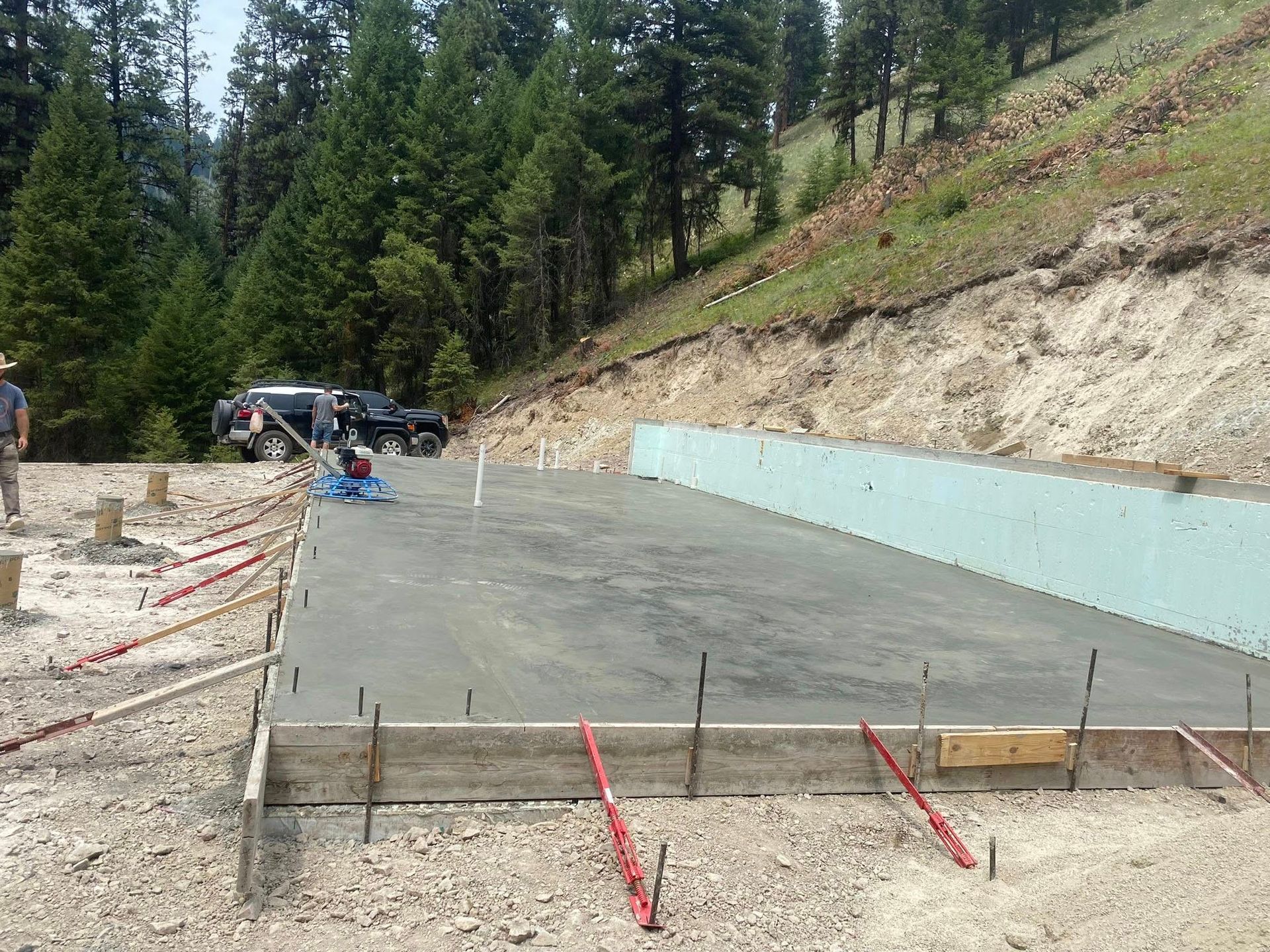 Concrete foundation slab being poured at a hillside construction site with workers and forest in the background