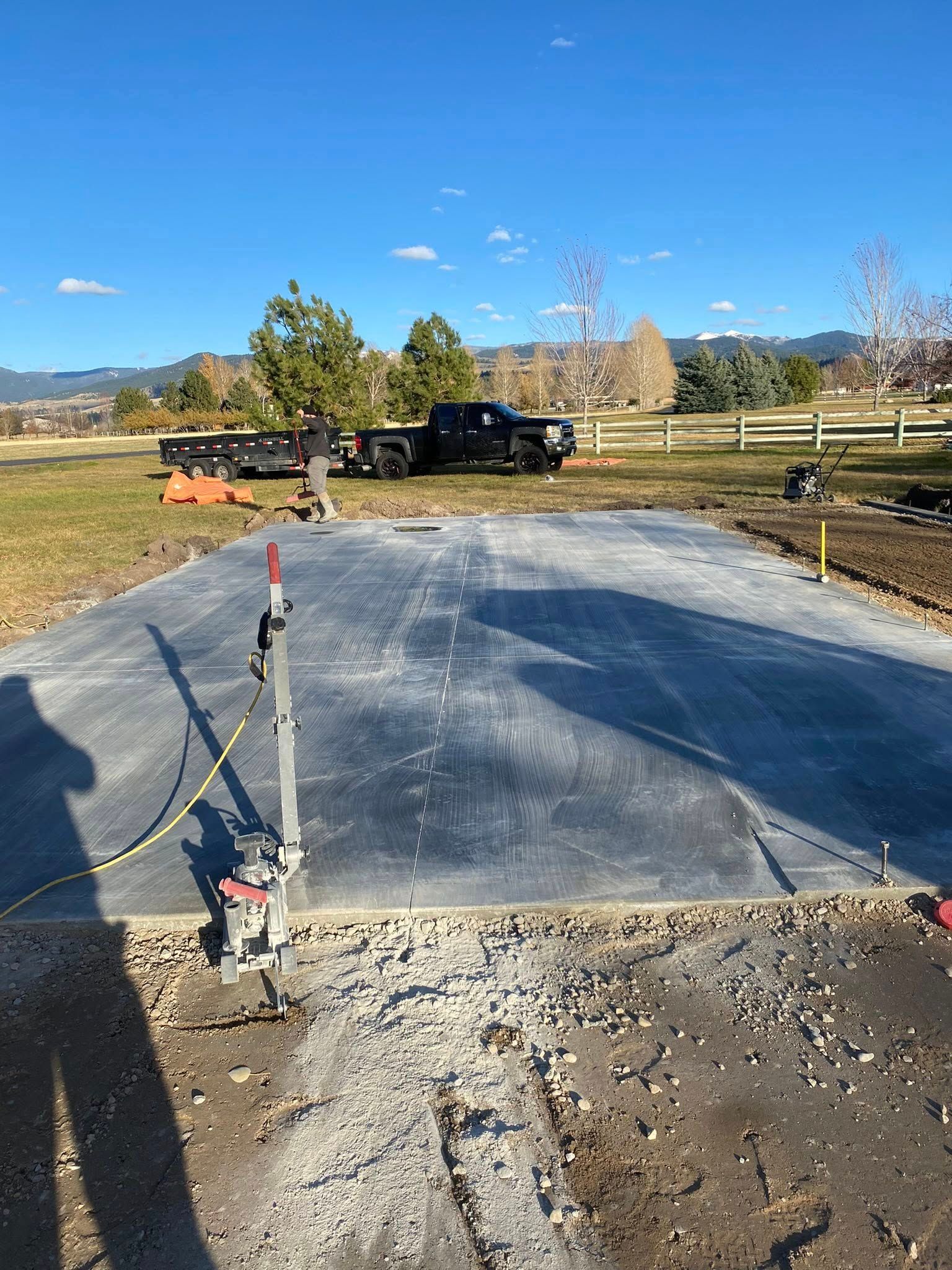 Freshly poured concrete pad under construction beside a road, with a fenced yard and truck in the background