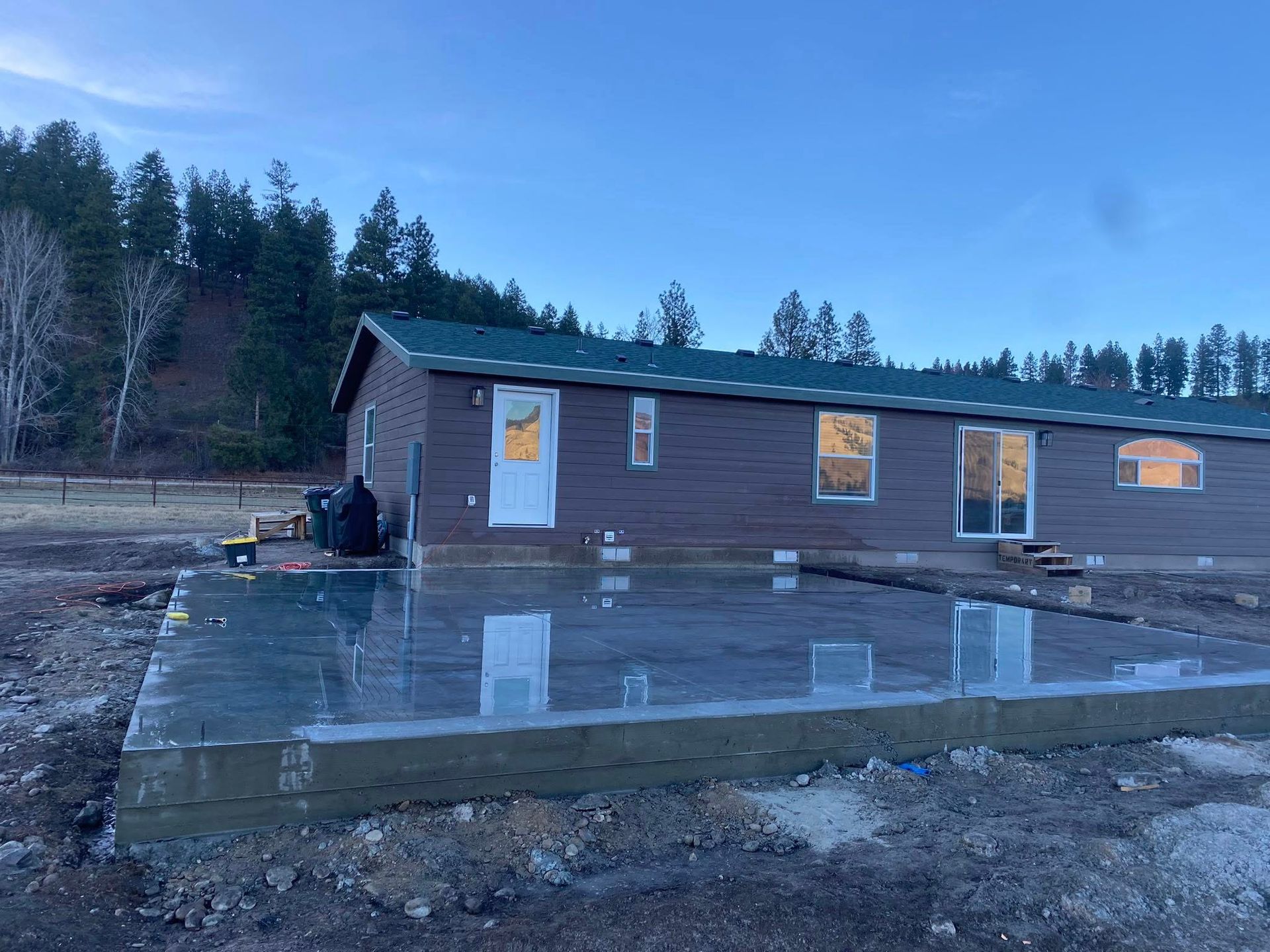 Small brown cabin on a muddy site with a wet concrete foundation, trees in the background.