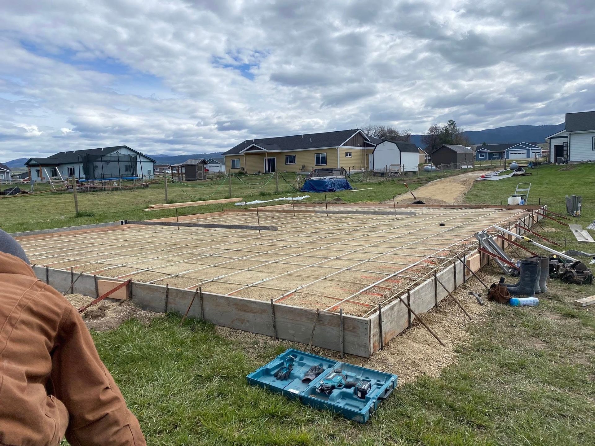 House foundation under construction in a grassy field, with wooden forms and scattered tools under cloudy skies