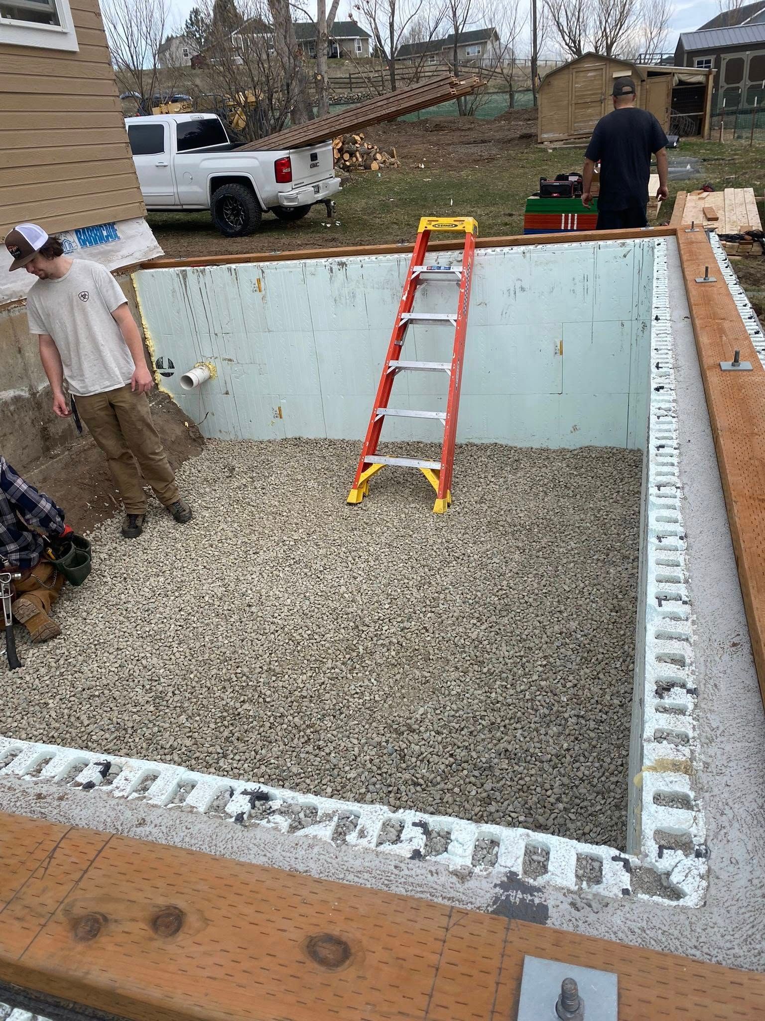 Partially dug backyard pool with a red ladder, gravel floor, and two people working around the edges
