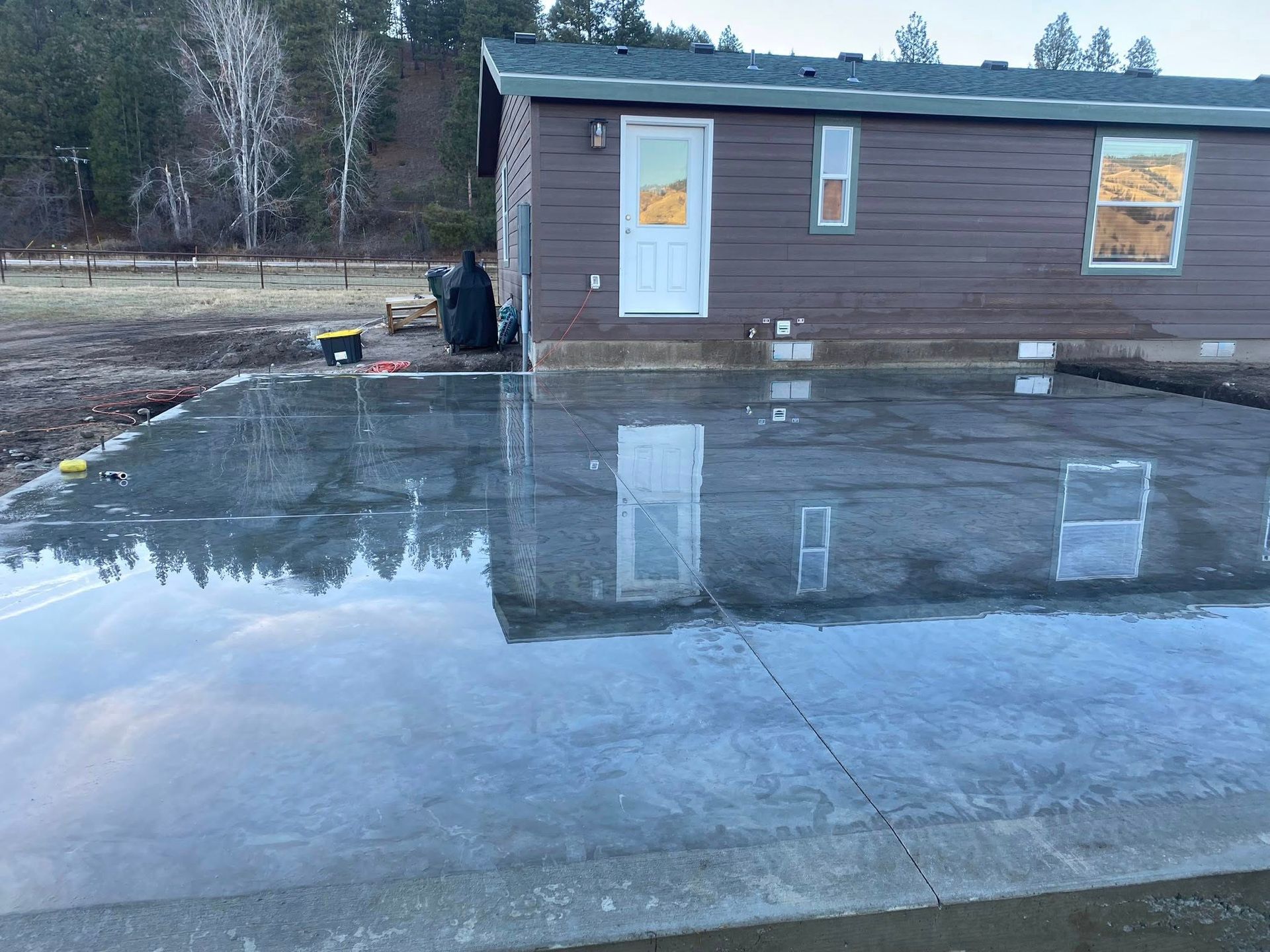Wet concrete driveway beside a brown cabin, reflecting the building and cloudy sky