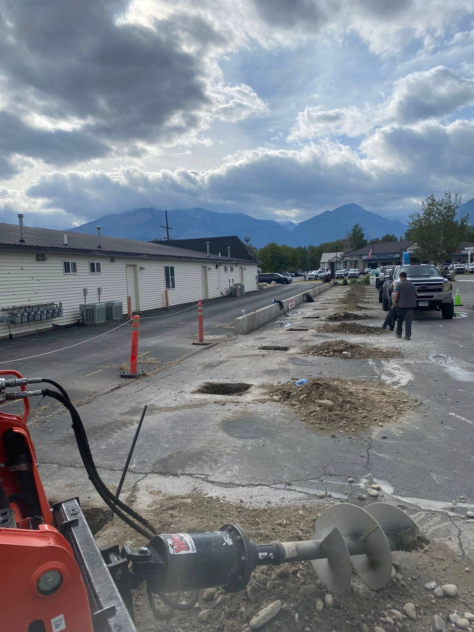 Construction site with excavator digging gravel beside a road under cloudy mountains.