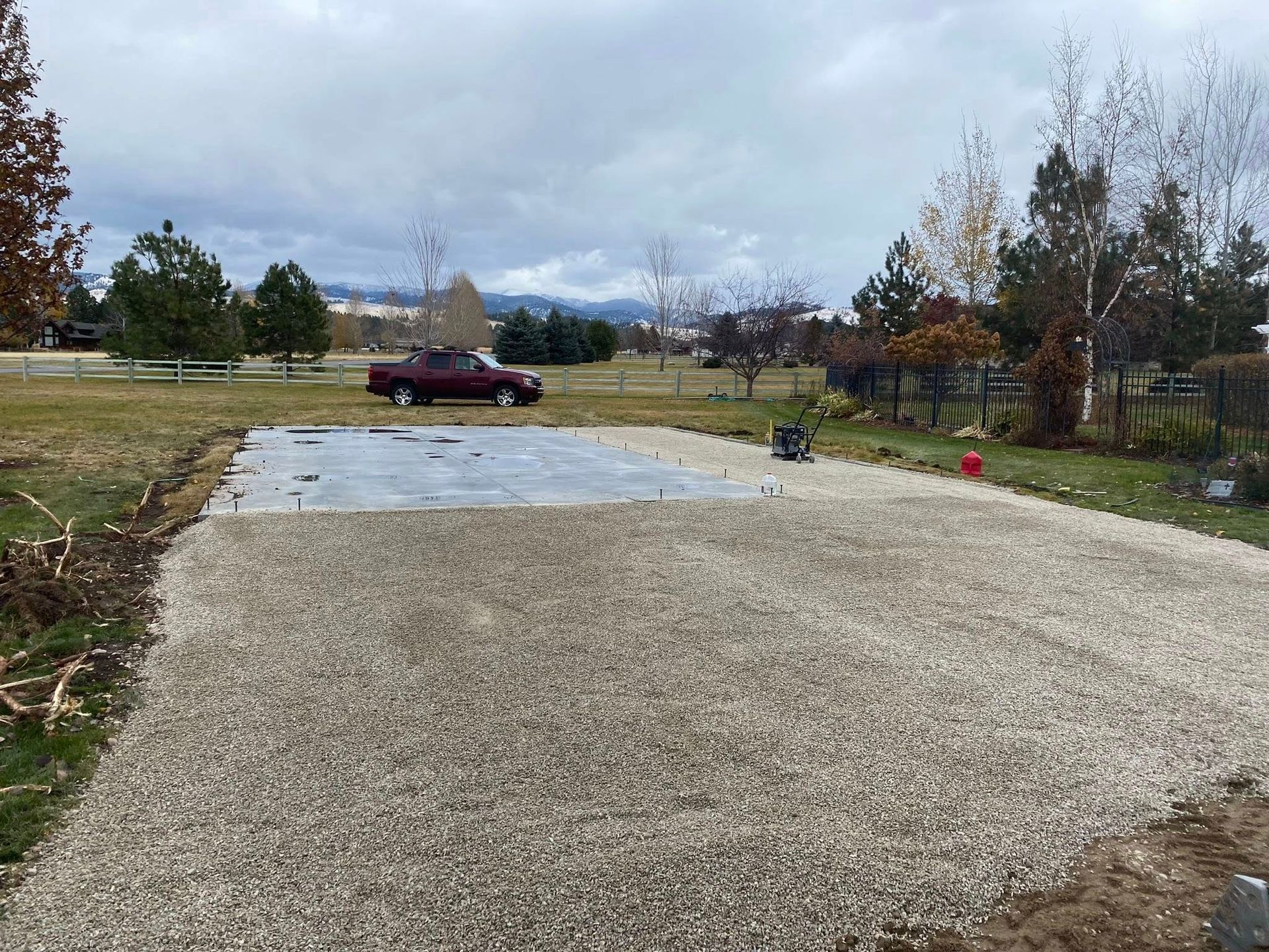 Gravel driveway under construction beside a parked maroon truck, with concrete slab and trees in the background