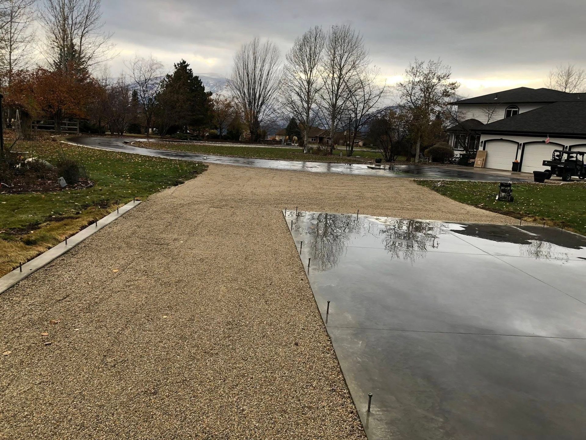 Gravel driveway with puddles beside a house and winter trees under a gray sky