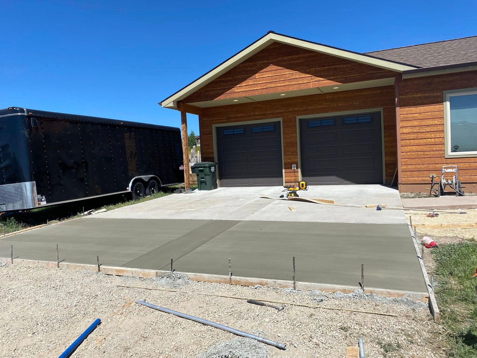 New concrete driveway in front of a brick garage with two black doors and a black trailer nearby