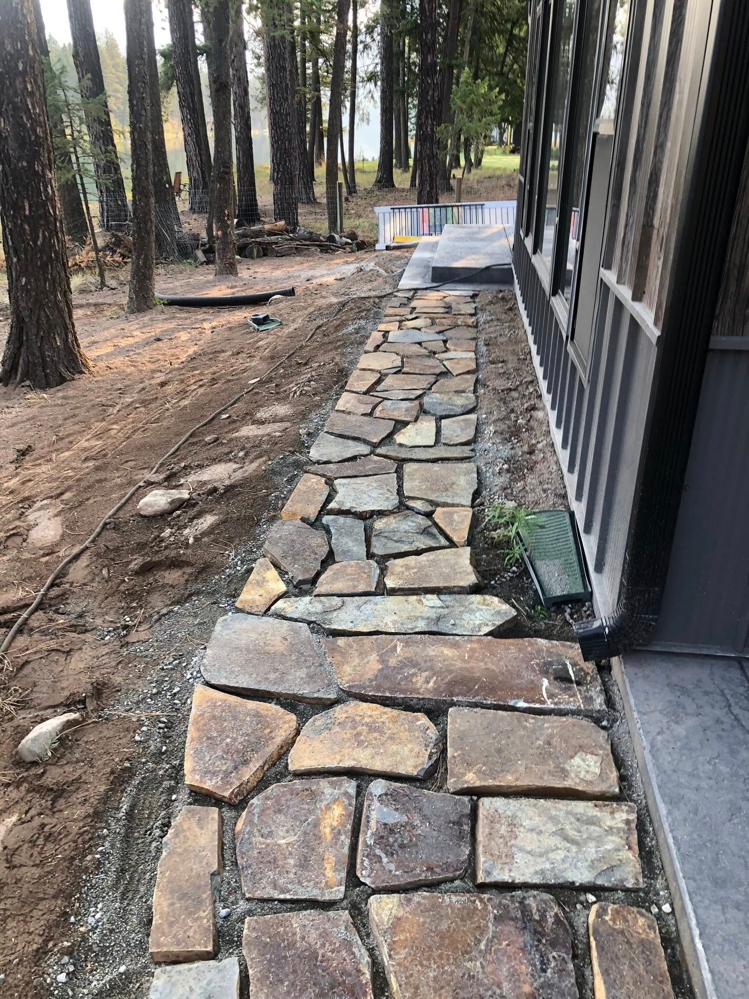 Stone walkway beside a black building, lined with trees and dirt ground.